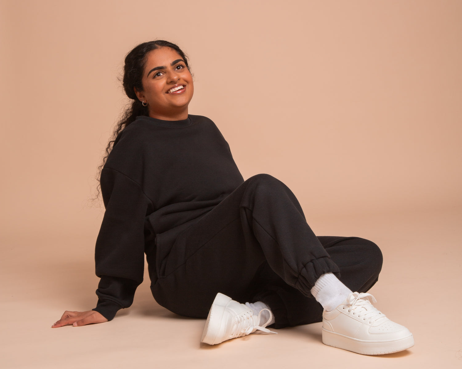 A person with long hair sits smiling on the floor, wearing a black tracksuit and white sneakers, against a beige background.