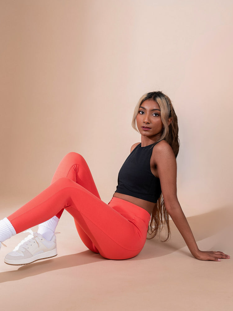 A person with long hair sits on a beige floor against a light background. They are wearing a black sleeveless top, bright orange leggings, white socks, and white sneakers, looking at the camera.