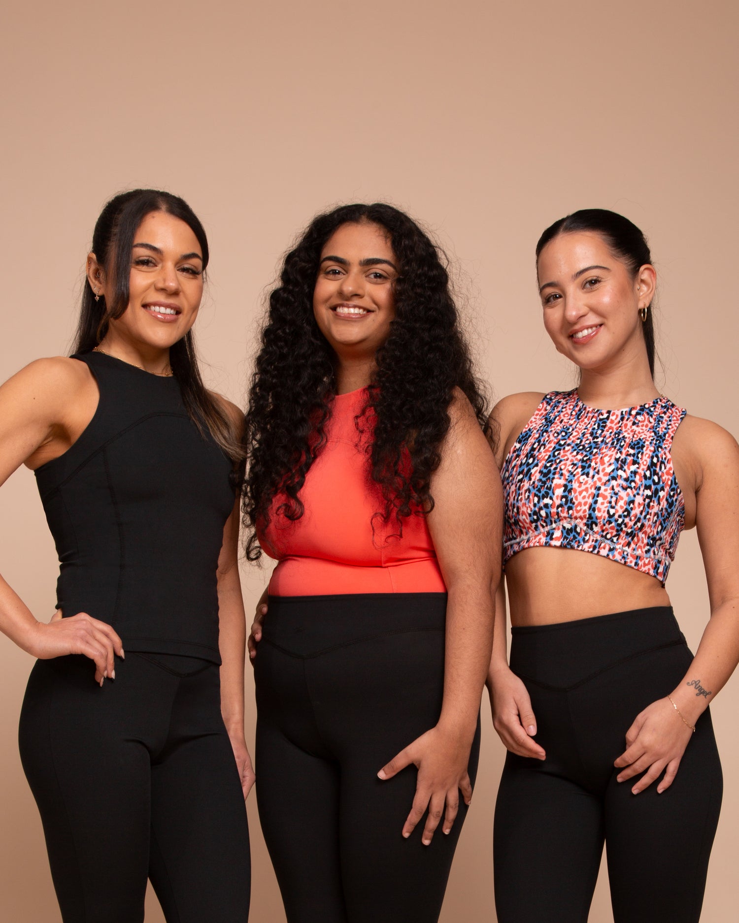 Three women in athletic wear stand together smiling against a neutral background. The first wears a black top, the second an orange top, and the third a colorful patterned top.