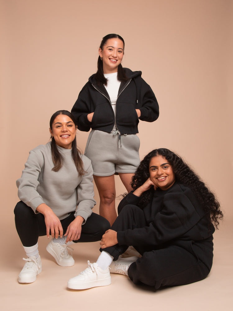 Three women in casual outfits pose against a beige background. One is standing, wearing a black hoodie and gray shorts, while the other two are kneeling, dressed in gray and black loungewear.