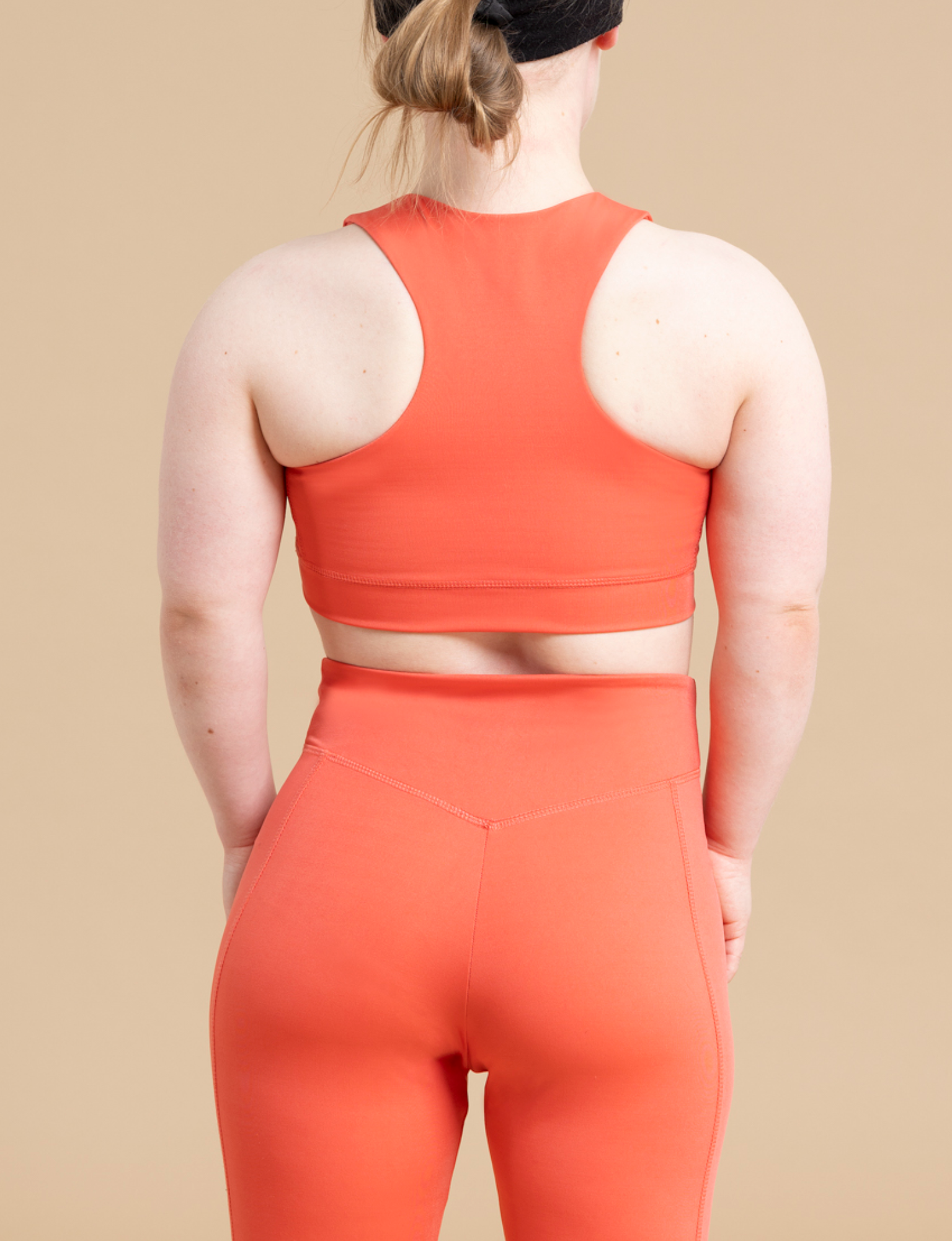 The image shows a person with light skin and blonde hair in a ponytail, viewed from the back. They are wearing an orange DEWEY HAZEL cropped vest top and matching high-waisted leggings, which blend perfectly against a beige background. This athleisure wear provides ideal light support for low-impact activities.