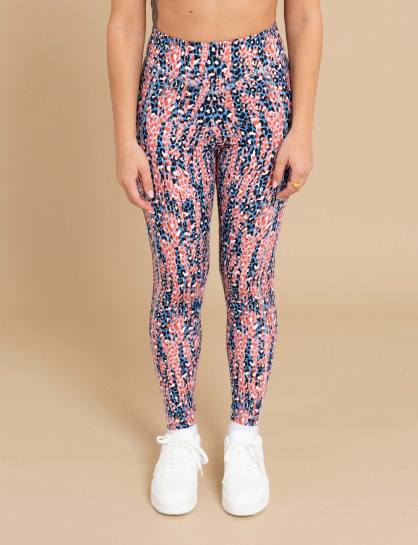 A person is standing against a beige background, wearing DEWEY's IRIS printed high waist leggings, featuring a mix of pink, blue, and black designs in an athleisure wear style. They are also wearing white sneakers. The photo captures them from the shoulders down with their arms resting by their sides.