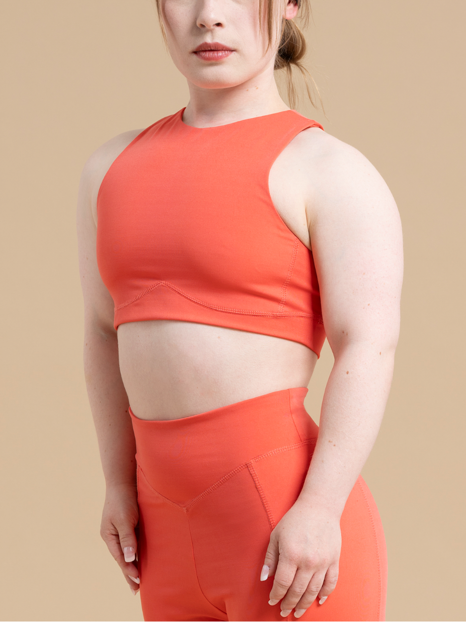 A person is wearing a matching orange athleisure outfit consisting of DEWEY's HAZEL cropped vest top and high-waisted leggings, offering light support. They stand against a plain beige background, their hair pulled back, gazing forward.