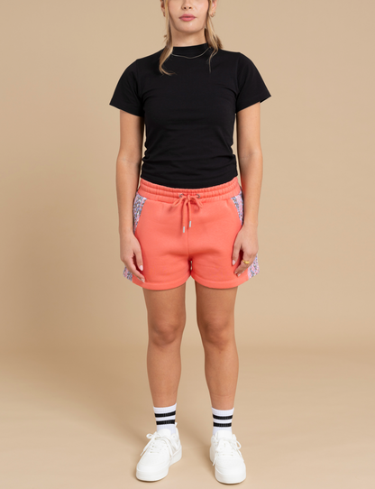 A person stands against a beige background wearing a plain black t-shirt, coral-colored DEWEY RILEY printed sweat shorts with patterned side panels, black and white striped socks, and white sneakers. Their hands rest by their sides, showcasing an elevated loungewear look.