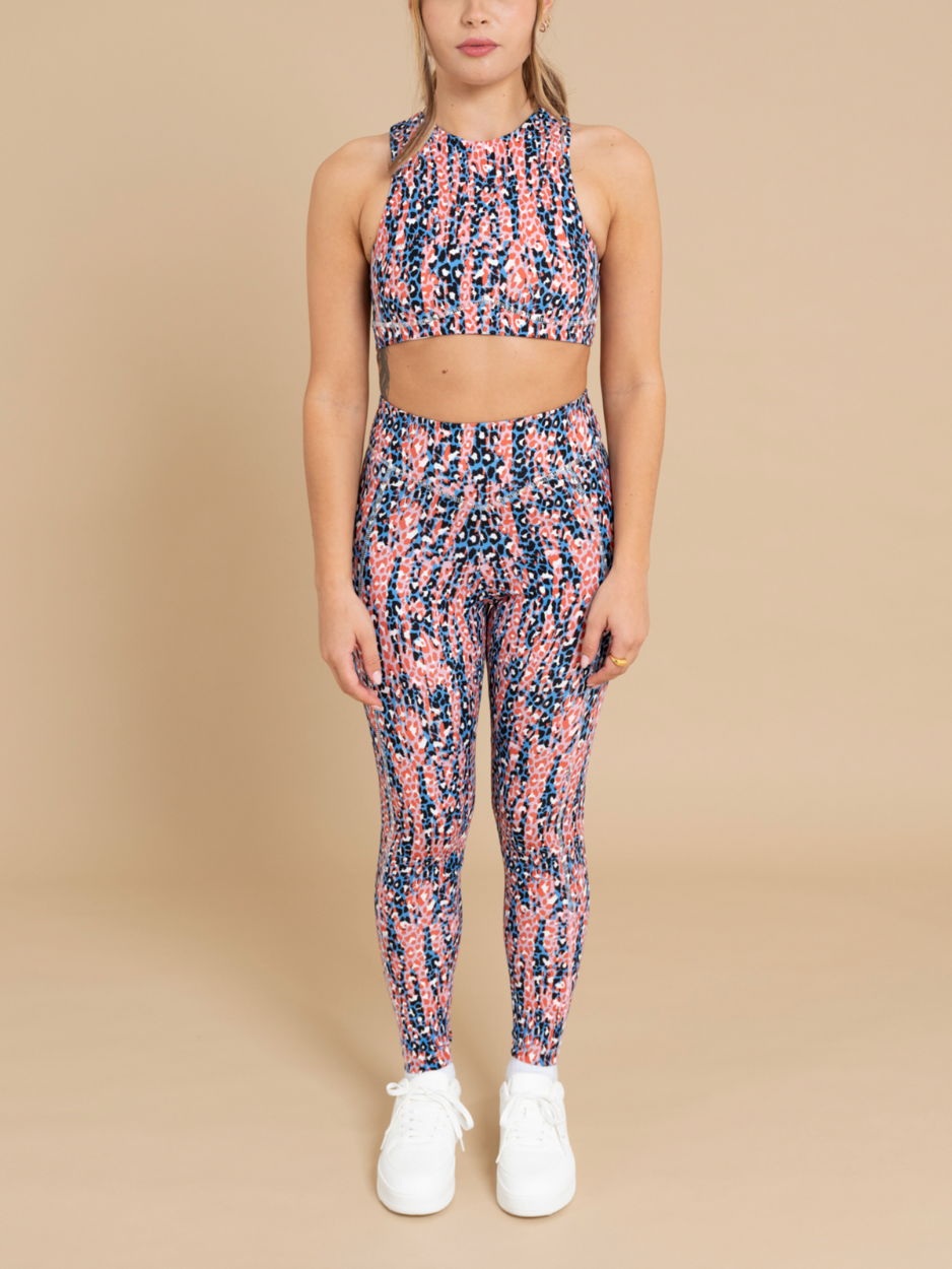 A person stands wearing a matching athleisure wear set with a colorful, abstract pattern in shades of pink, blue, and orange. The outfit includes a sleeveless crop top and DEWEY's IRIS printed high waist leggings that accentuate contour lines. The individual is also wearing white sneakers and stands against a beige background.