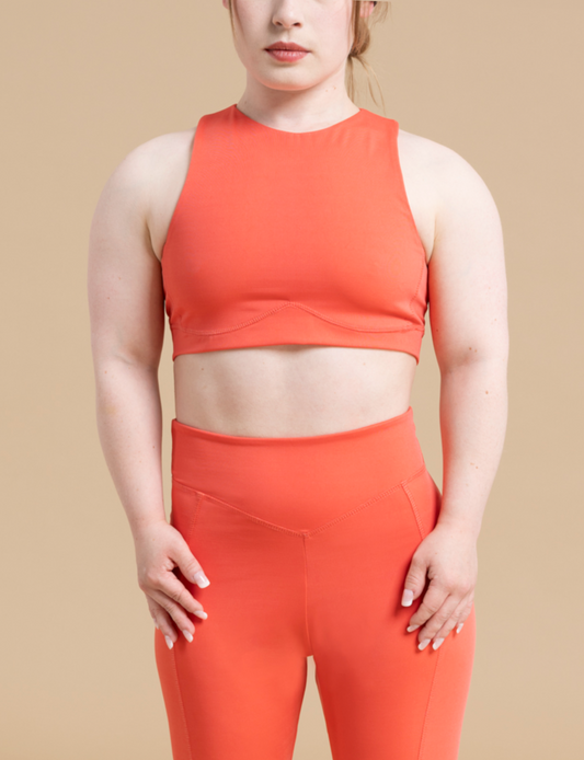 A person stands against a beige background wearing a matching orange DEWEY HAZEL cropped vest top and high-waisted leggings. Their arms are at their sides, with their thumbs tucked into the waistband of the leggings. They are looking straight ahead, showcasing the stylish athleisure wear.