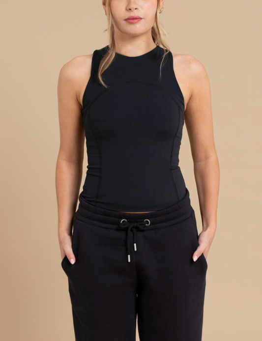 A person wearing a black, sleeveless, high-neck top and black drawstring pants stands against a beige background. Their hair is tied back, hands in pockets, exuding a casual and relaxed vibe. The SUMMER racer-back vest top by DEWEY adds a touch of sporty athleisure to the overall look.