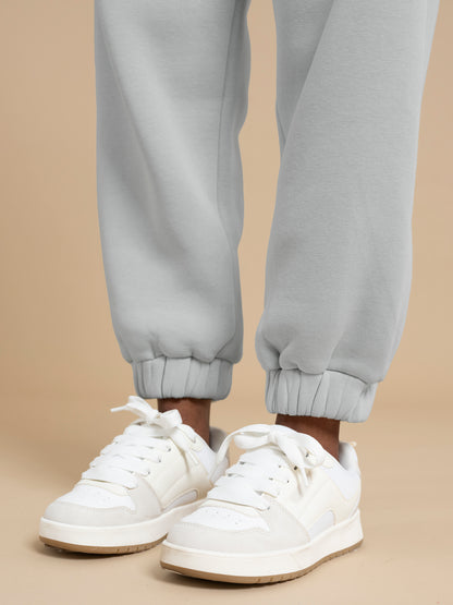 Close-up of a person with an extra-petite frame wearing white sneakers with laces and light gray jogger-style ADA sweatpants by DEWEY. The pants feature elasticated cuffs, set against a soft beige background, showcasing the lower part of their legs.