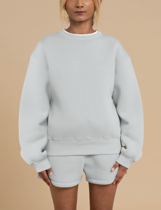 A person standing against a beige background is wearing a light grey oversized DEWEY BAILEY crew neck sweater with matching shorts. Their hands rest relaxed by their sides, highlighting the ensemble's perfect fit. With their hair tied back, the harmony of style and comfort is accentuated.