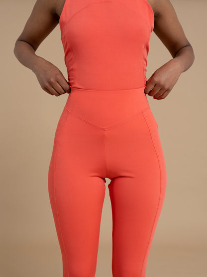 A person sporting a coral-colored athletic outfit adjusts their figure-flattering IRIS high waist leggings by DEWEY. The soft and stretchy fabric enhances comfort against the solid beige background.