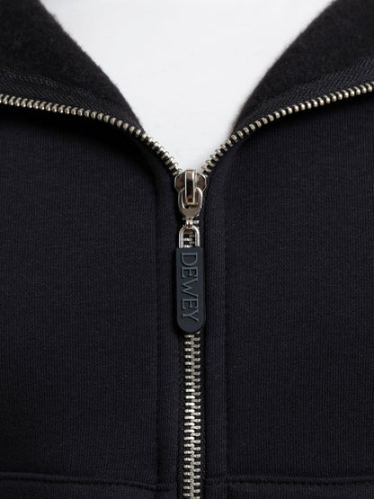 Close-up image of a black DEWEY TOBY full zip hoodie. The zipper is partially closed, with the zipper pull tab labeled "DEWEY." The fabric around the zipper appears to be a soft, black material, giving it an athleisure look.