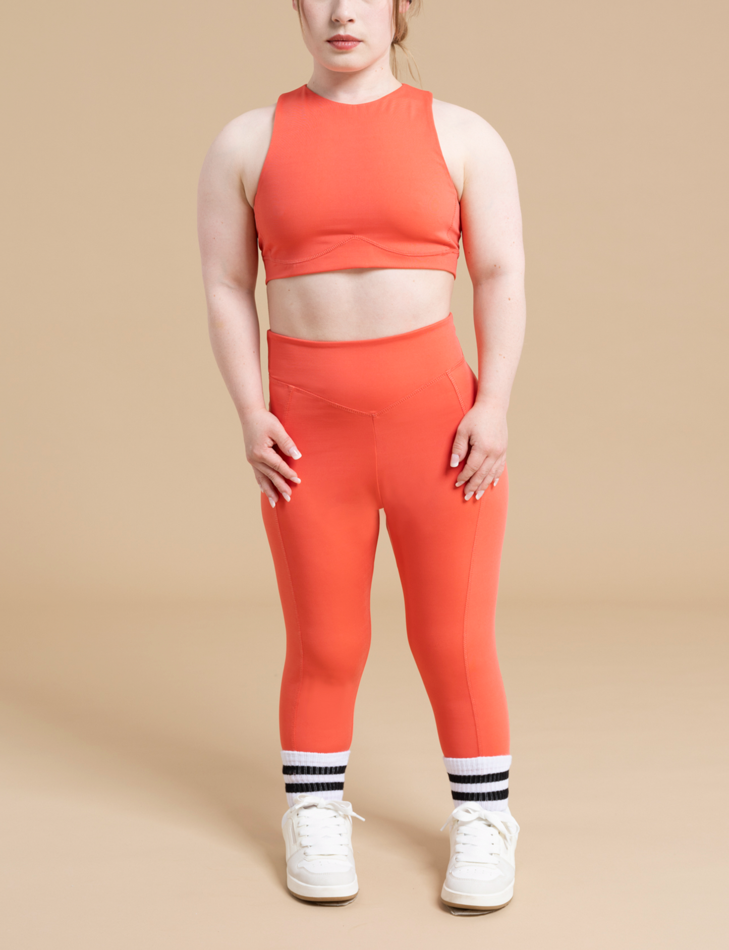 A person with long auburn hair is posing against a beige background, wearing the DEWEY HAZEL cropped vest top in orange and matching high-waisted leggings. They are also wearing white sneakers and white athletic socks with black stripes, creating a perfect athleisure wear look with light support.