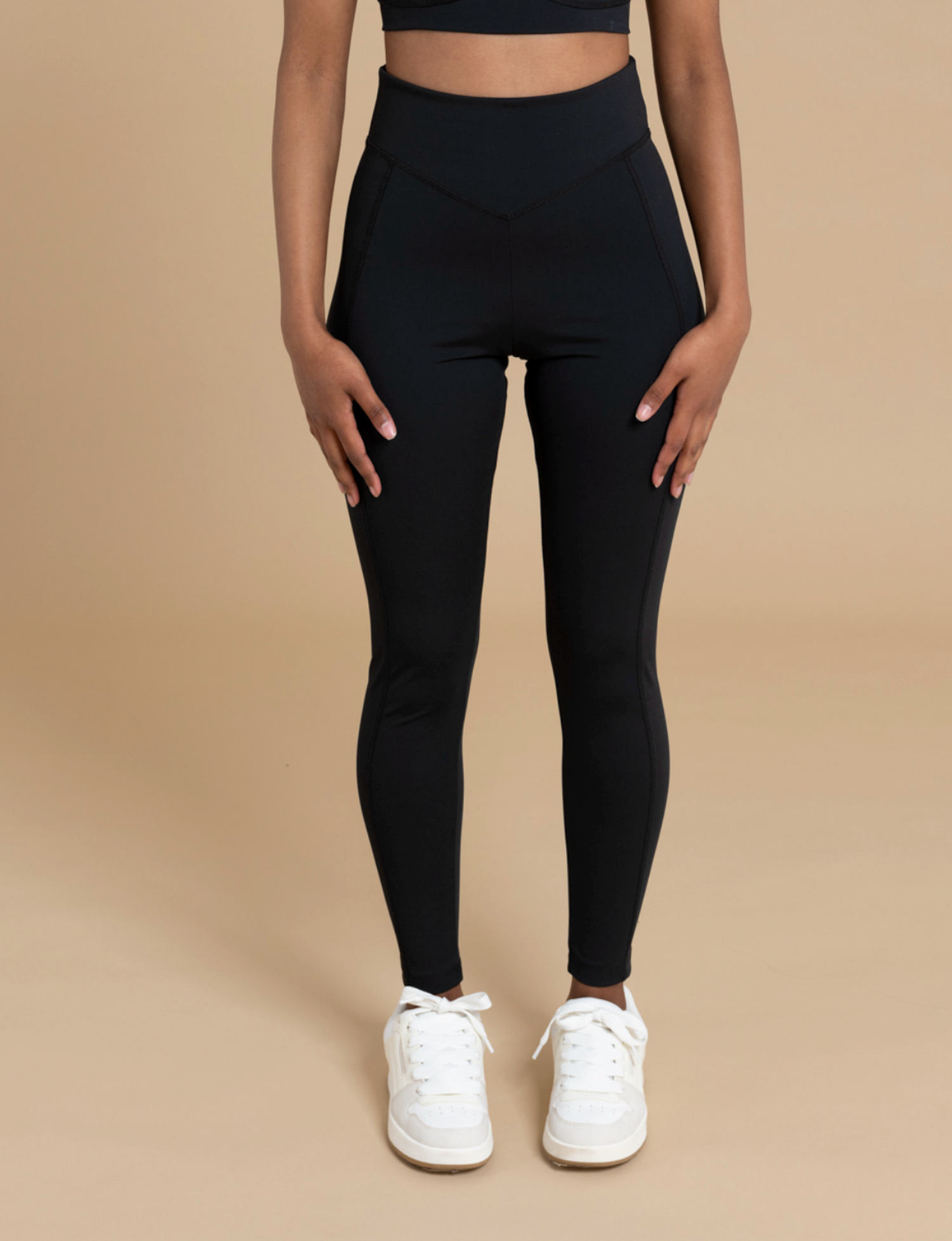 A person wearing DEWEY's IRIS high-waist leggings in a soft and stretchy fabric, paired with white sneakers, stands against a neutral beige background. Their upper body is largely cropped out of the image, emphasizing the flattering silhouette of the leggings.