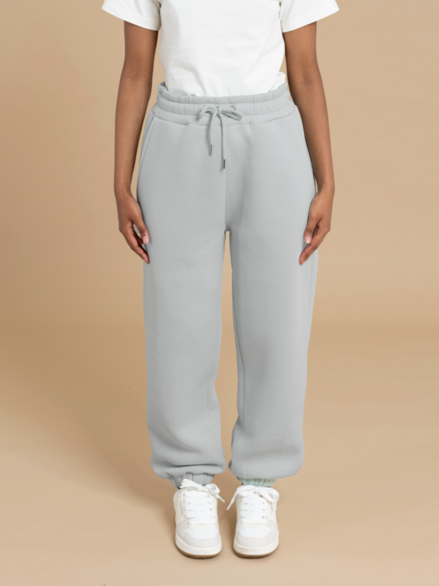 A person is standing against a beige background, dressed in a white t-shirt, light gray DEWEY ADA sweatpants with a drawstring, and white sneakers. The focus is on the lower half of the body, highlighting the casual and comfortable outfit that is perfect for an extra-petite frame.