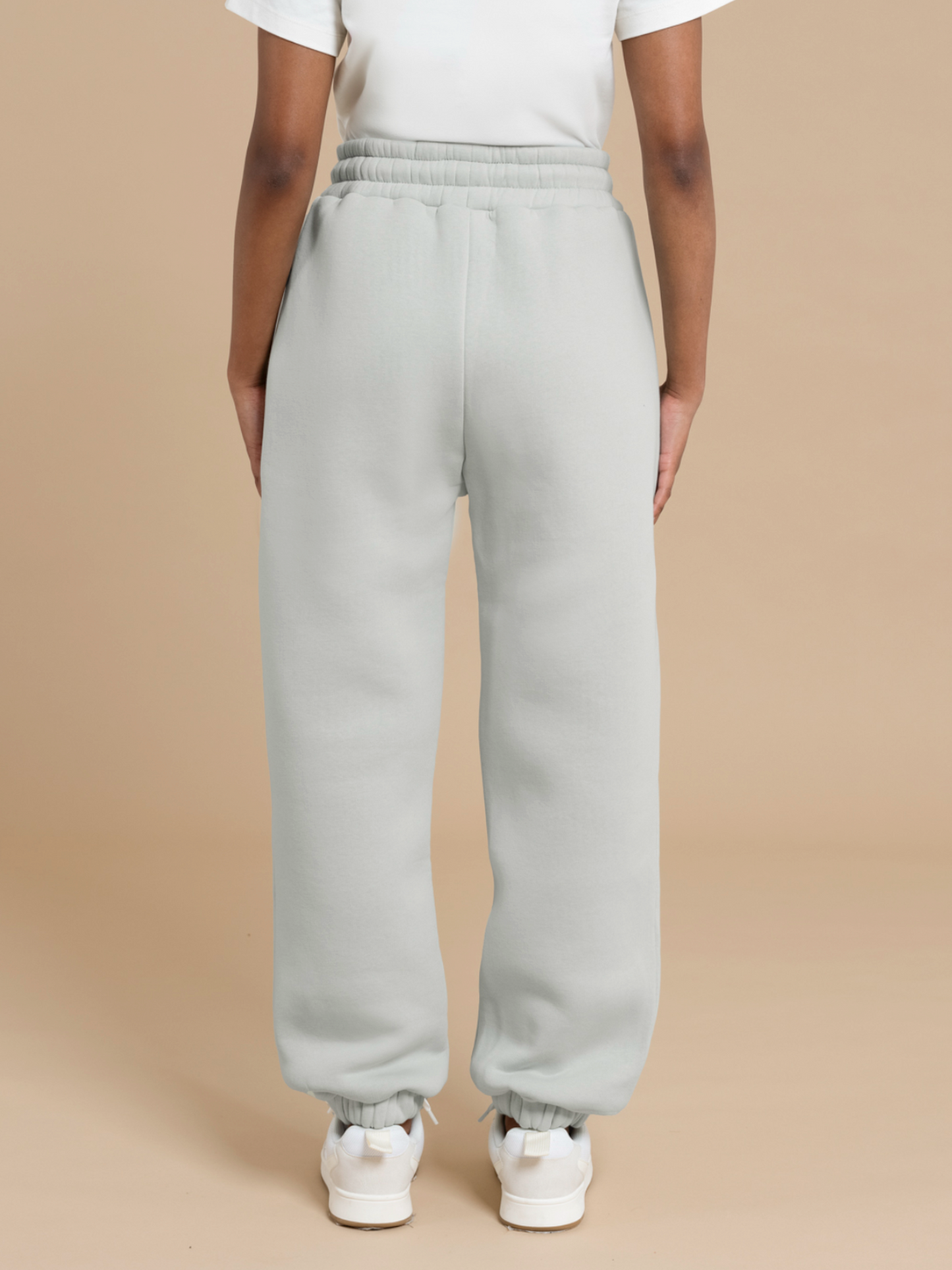 A person with an extra-petite frame is standing with their back to the camera, showcasing light grey ADA sweatpants from DEWEY. The sweatpants feature an elastic waistband and cuffs and are paired with white sneakers. The background is a plain beige color.