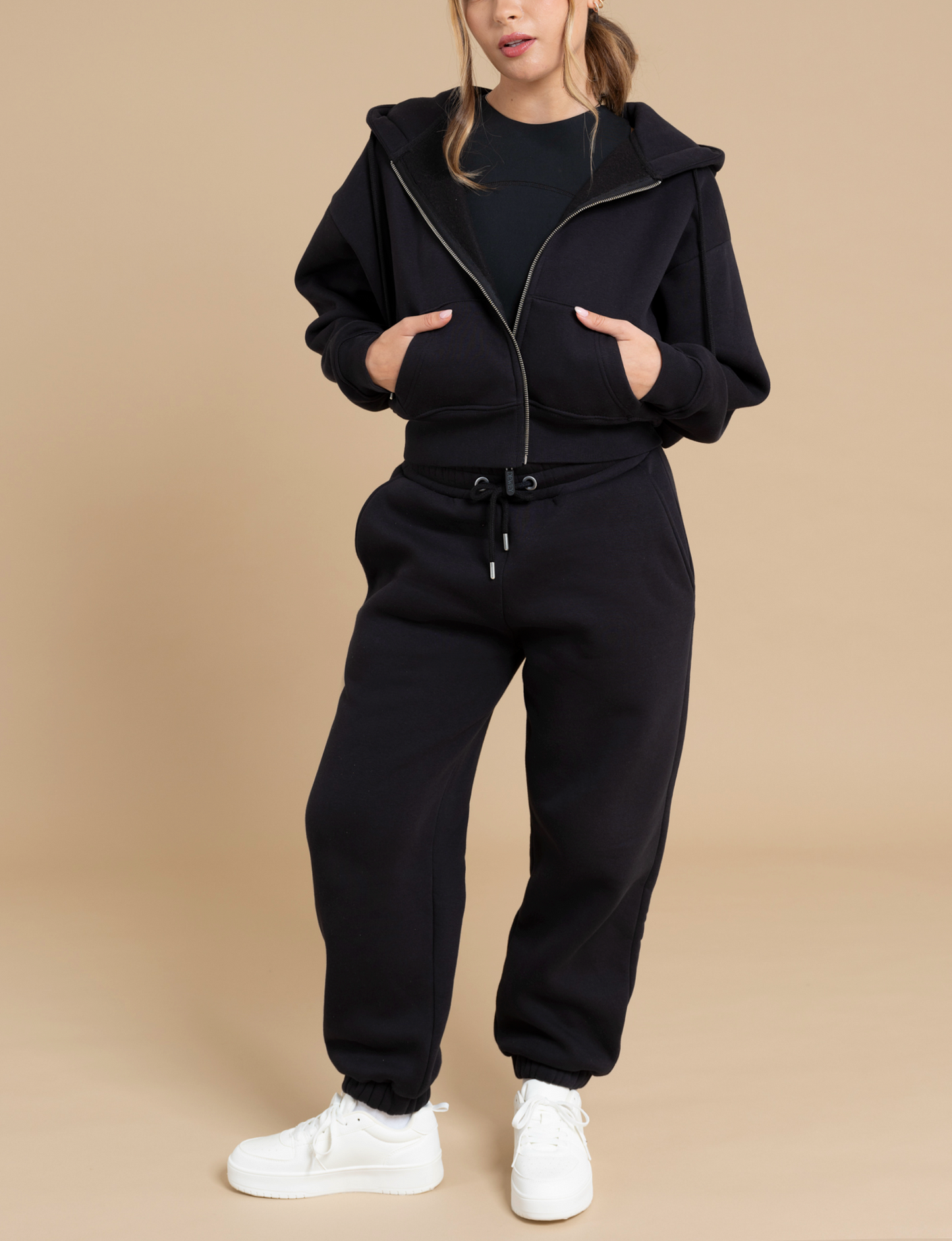 Against a plain beige background, a person models an effortless athleisure look by wearing a black DEWEY TOBY full zip hoodie paired with matching black sweatpants and white sneakers. Their hair is partially tied back, and they pose with one hand in a pocket while the other rests on their hip.