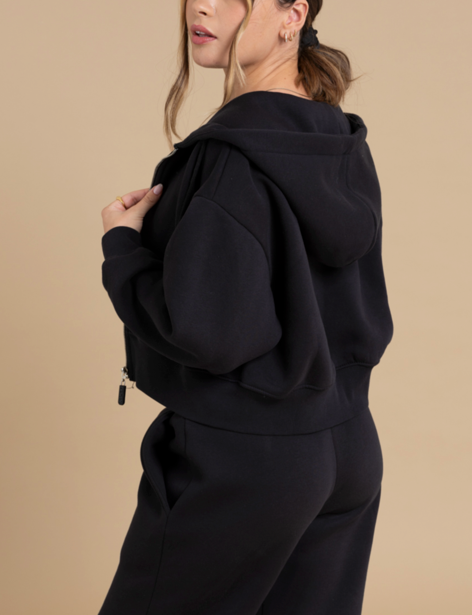 A person standing against a beige background is wearing a black TOBY full zip hoodie from DEWEY and black pants. Their hair is tied back, and they are looking slightly over their shoulder. The hoodie, featuring a zipper and hood, offers a specialized fit that enhances the casual, athleisure look of the outfit.