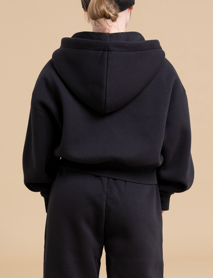 A person wearing a black TOBY full zip hoodie by DEWEY and black pants stands facing away from the camera against a beige background. The specialized fit hoodie is oversized with the hood up, covering the person's head and ears, creating an athleisure look. Their hair is not visible.