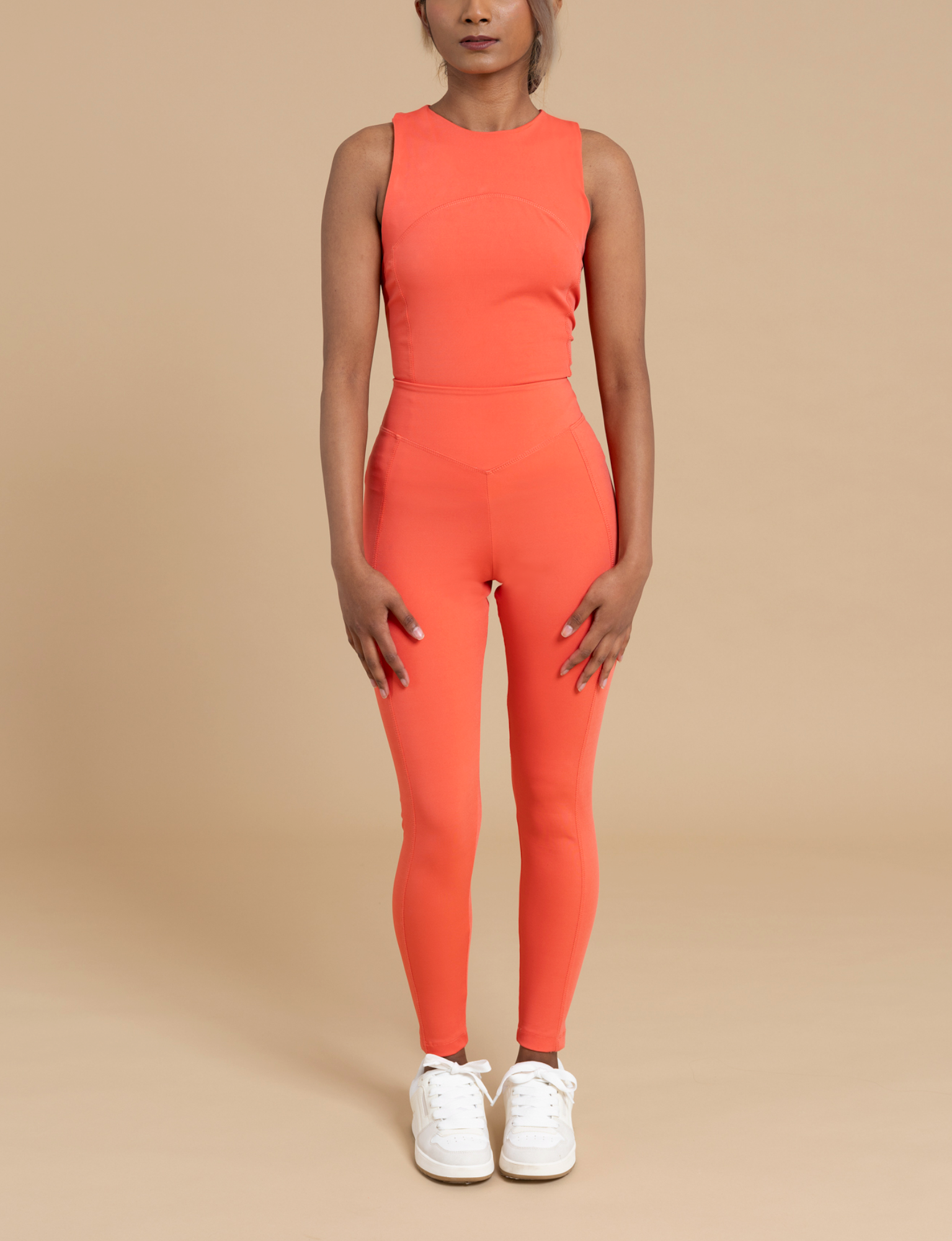 A person stands facing forward wearing a sleeveless, fitted, bright orange athletic top and matching IRIS high-waist leggings from DEWEY. They have their arms relaxed by their sides and are wearing white sneakers. The soft and stretchy fabric highlights a flattering silhouette against the plain beige background.