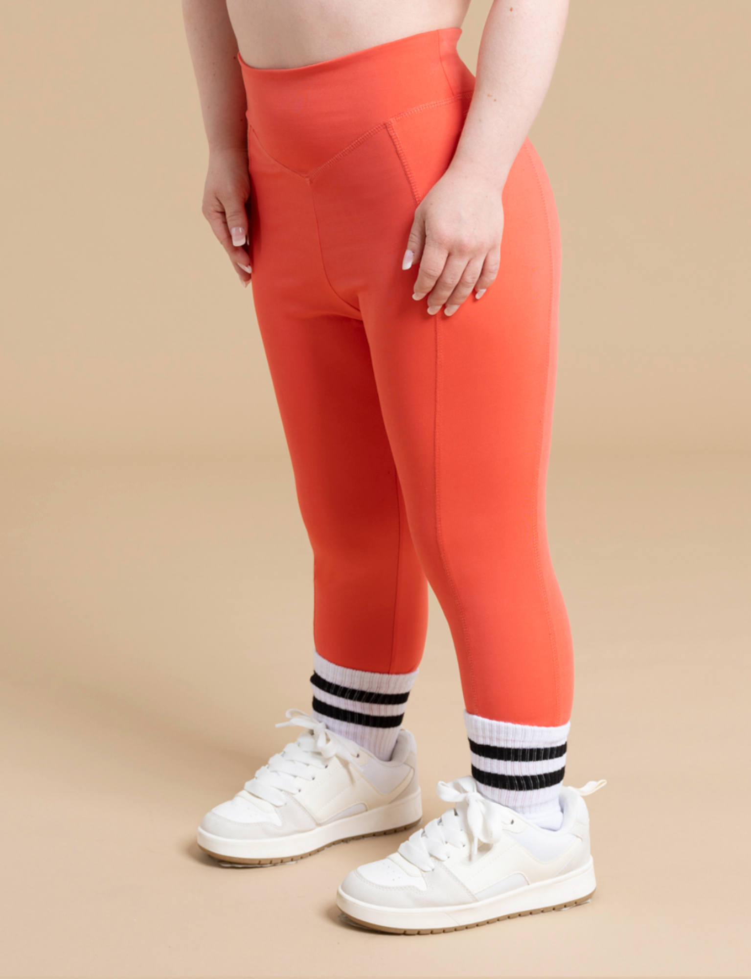 A person wearing DEWEY's IRIS high-waist leggings in bright orange, paired with striped socks and white sneakers, stands against a beige background. These leggings not only offer a flattering silhouette but are also crafted from soft and stretchy fabric for ultimate comfort. The view is from the person's chest down.