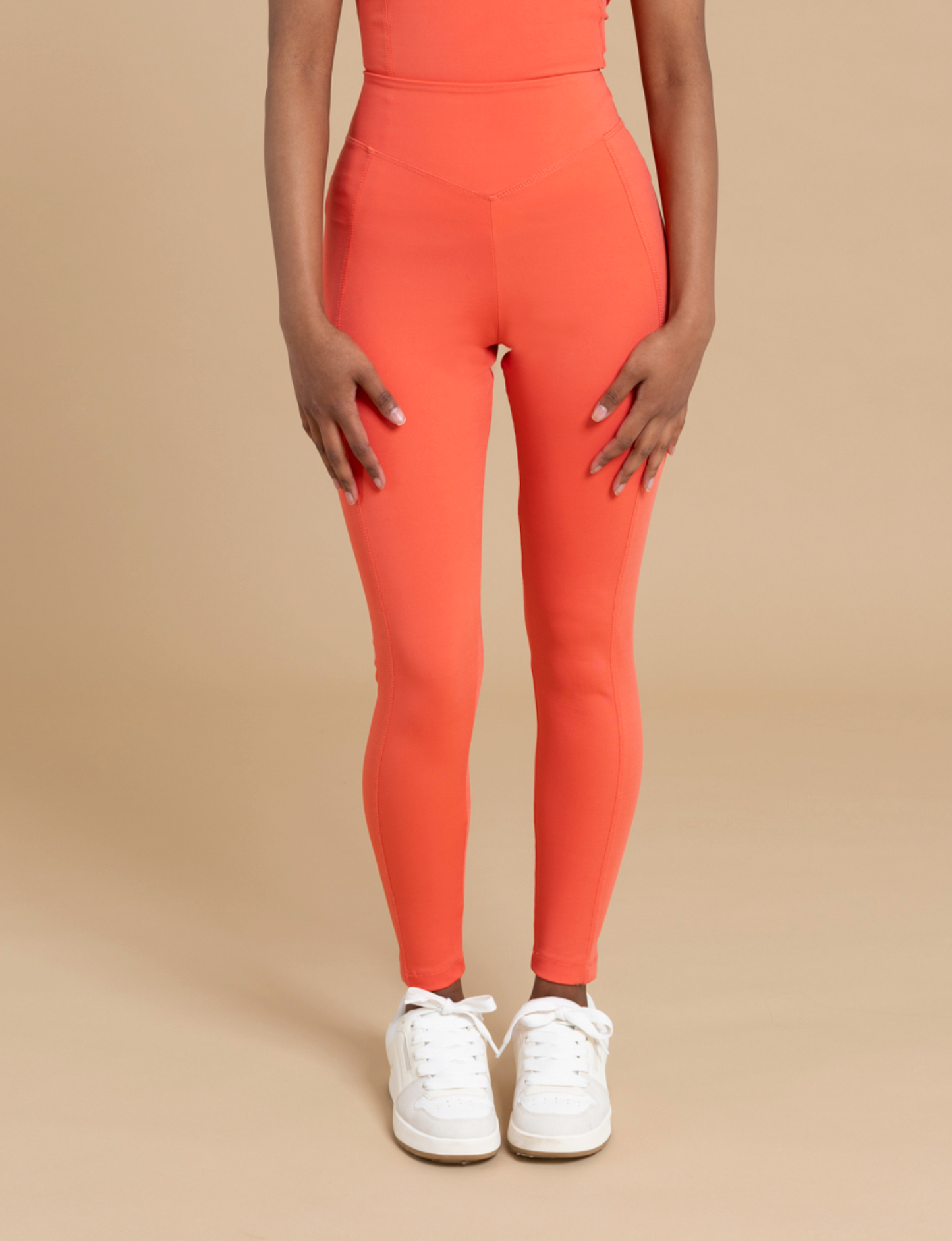 Standing against a beige background, a person is seen wearing DEWEY's IRIS high-waist leggings in a vibrant orange. These form-fitting leggings create a flattering silhouette and pair perfectly with white sneakers. Their hands rest relaxed by their sides, while their upper body is outside the frame.