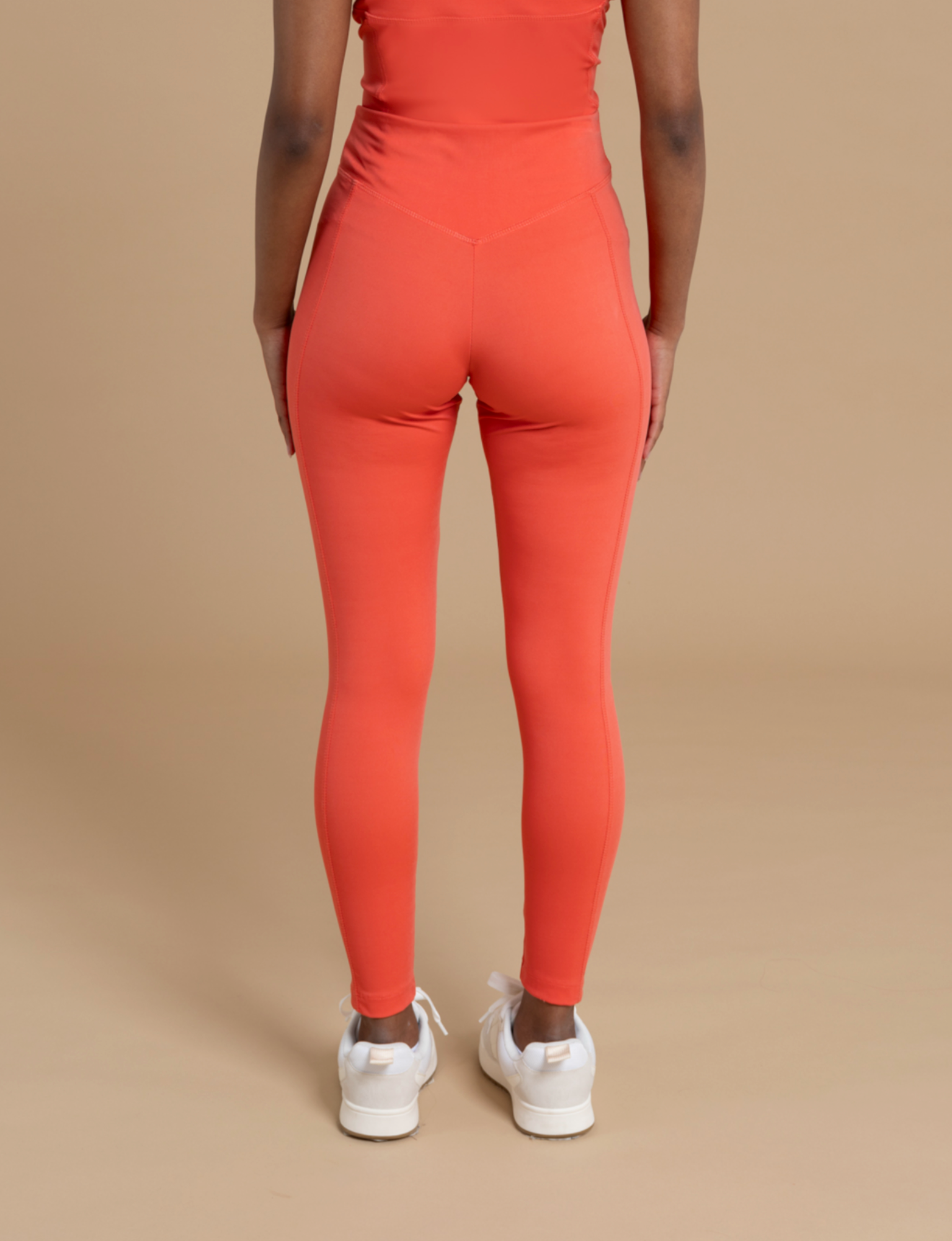 The image features a person standing with their back to the camera, beautifully highlighting a flattering silhouette in DEWEY's IRIS high-waist leggings in bright orange. They are paired with a matching orange top and white sneakers, all set against a plain beige background. The soft and stretchy fabric of the leggings guarantees maximum comfort.