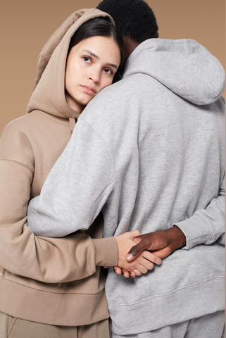 A woman in a beige hoodie embraces a man in a gray hoodie from behind, resting her head on his shoulder. Their hands are gently clasped together. The background is a simple, warm shade of brown.