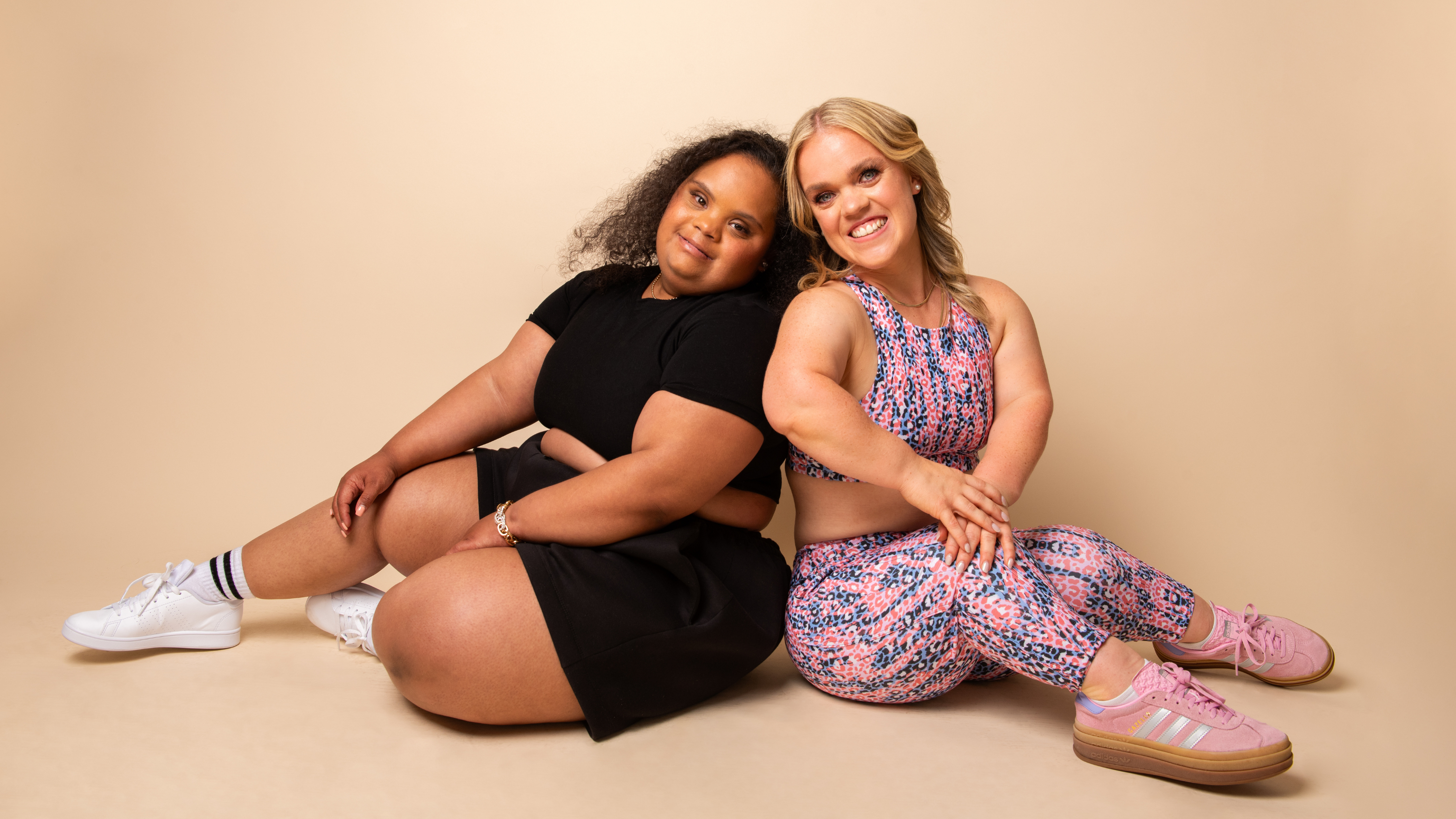 Two women sitting on the floor against a beige background, smiling. The woman on the left has dark curly hair, wears a black dress, white sneakers, and striped socks. The woman on the right has blonde hair, wears a colorful sports outfit with pink sneakers.